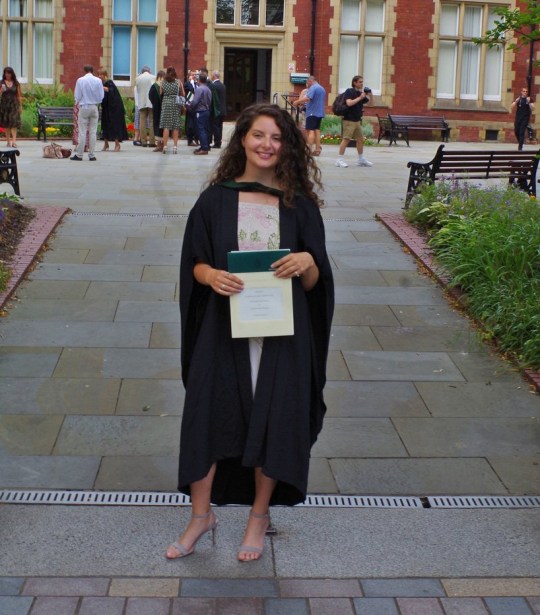 Chloe on her graduation day, wearing her graduation gown, the heels Dean asked her​ about on Tinder and holding a document, with ⁤a red brick building‍ behind her